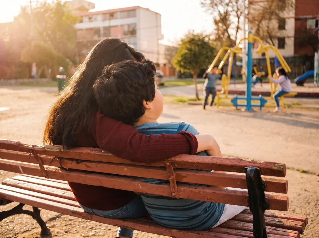Una mujer y un niño sentados en un banco frente a una plaza de juegos